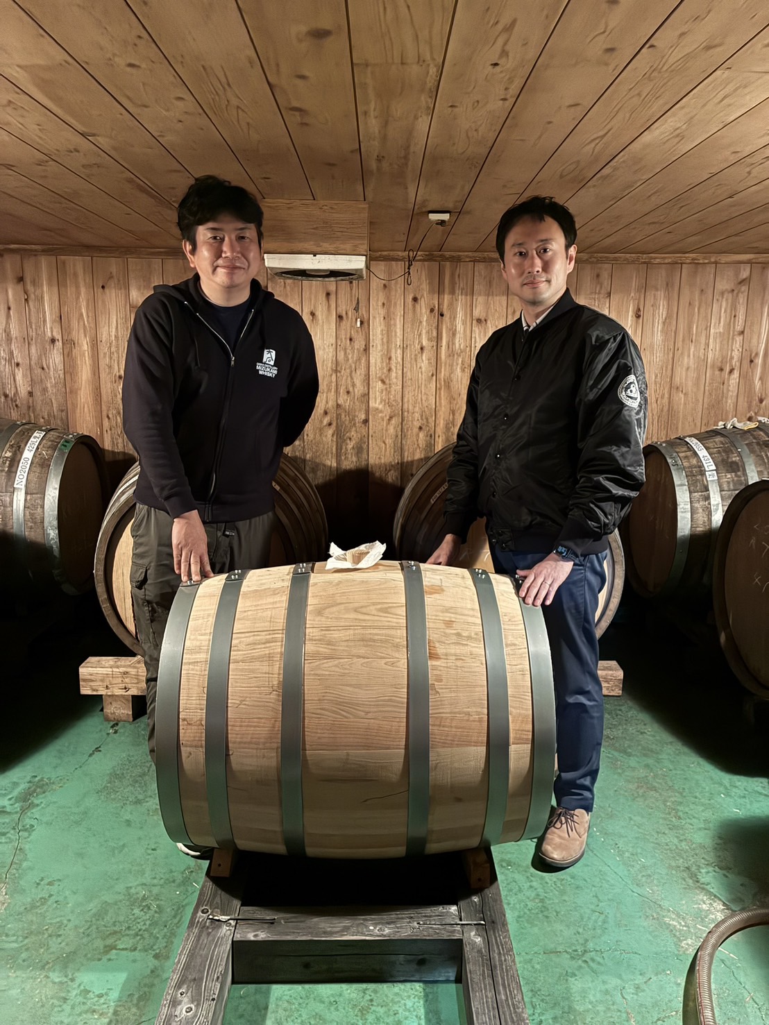 木造の貯蔵庫で、サクラ樽を前に並ぶ二人。サクラ樽に原酒を詰めた日の写真。